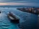An unbranded oil tanker navigating a narrow shipping channel beside refinery storage tanks at dawn.