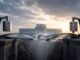 Two mechanical counterweights balance an abstract steel-ribbon bridge in front of a central-bank building at sunrise.